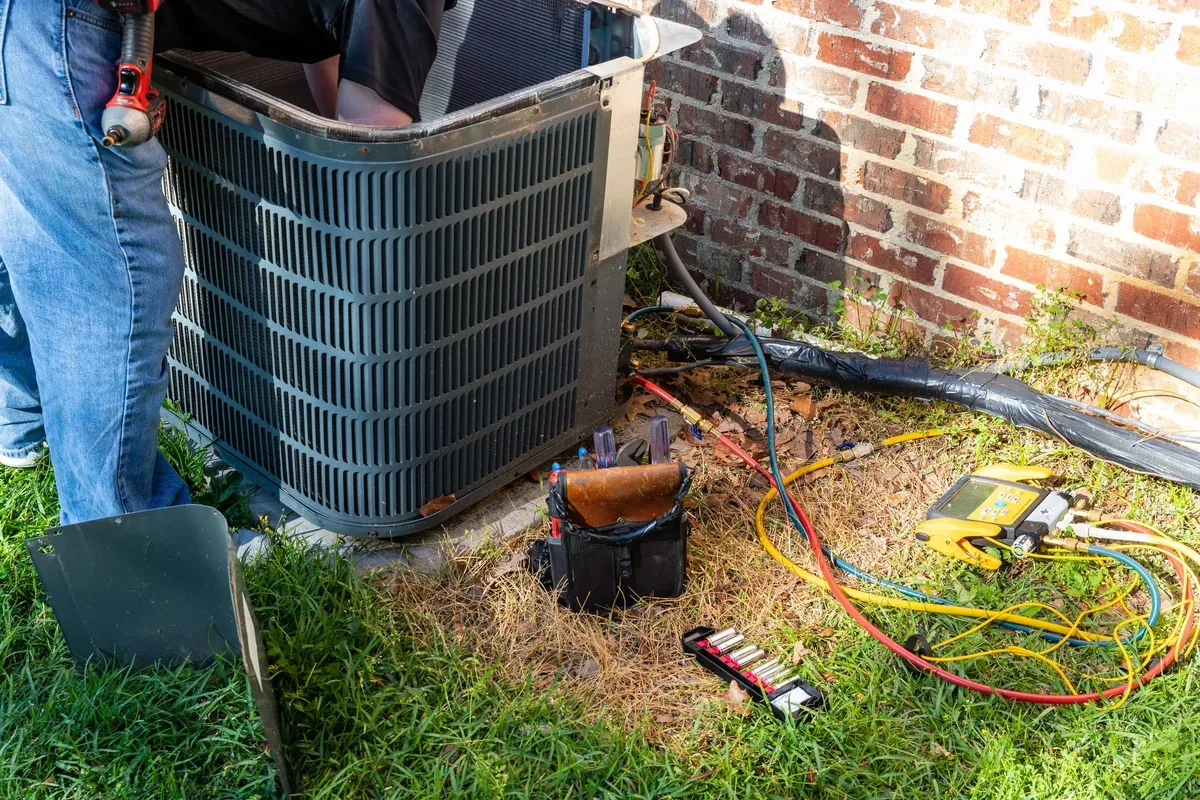 HVAC technician servicing an outdoor air conditioning condenser unit beside a brick home